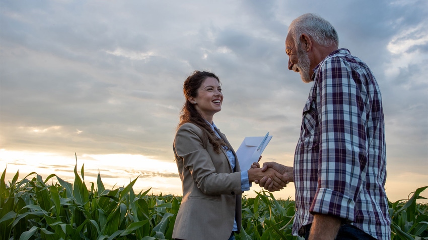 A senior farmer shaking hands with young woman with notebook in cornfield