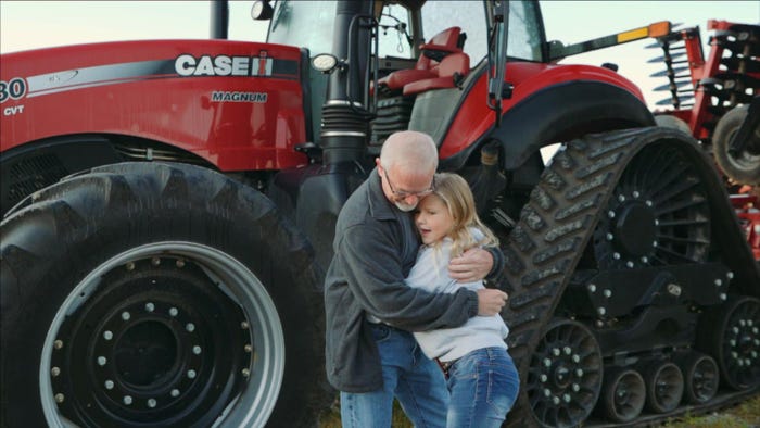 A man and a young girl embrace in a hug while standing in front of a red tractor