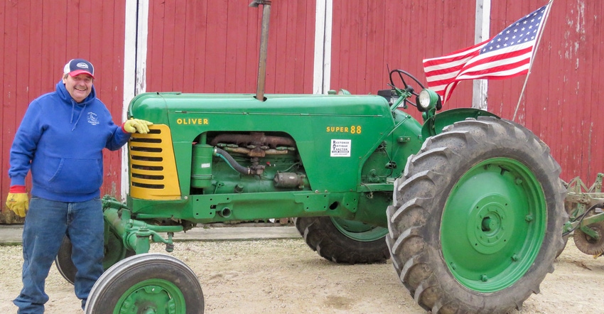 A man in a blue sweatshirt leaning against a green tractor with an American flag attached to it A man in a blue sweatshirt leaning against a green tractor with an American flag attached to it
