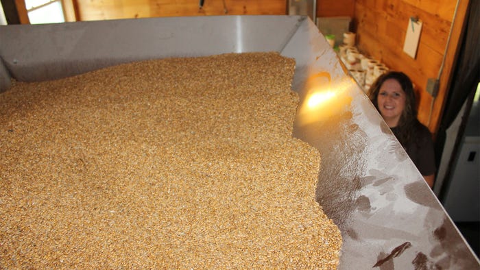 A large metal container filled with wheat berries and a woman smiling in the background A large metal container filled with wheat berries and a woman smiling in the background