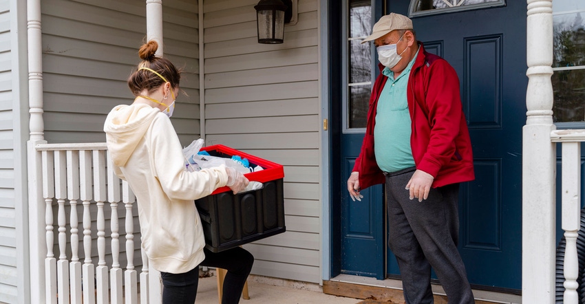 A young woman wearing a protective mask delivers groceries to a senior man on his porch during the COVID-19 outbreak