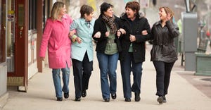 Group of women walking down sidewalk arm-in-arm in small town.