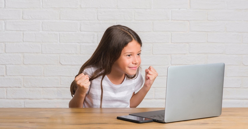 A young girl sitting at a table with a laptop and an excited expression