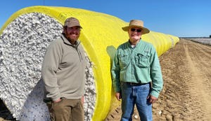 Two men stand by bales of cotton.