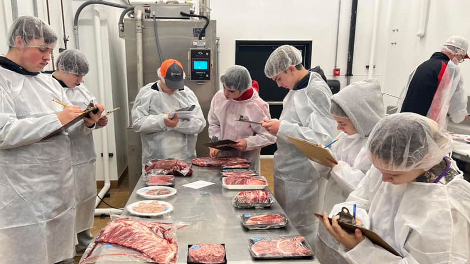 College students dressed in butcher coats and hair nets standing around a table, holding clipboards and rating butcher cuts of meat on the table.