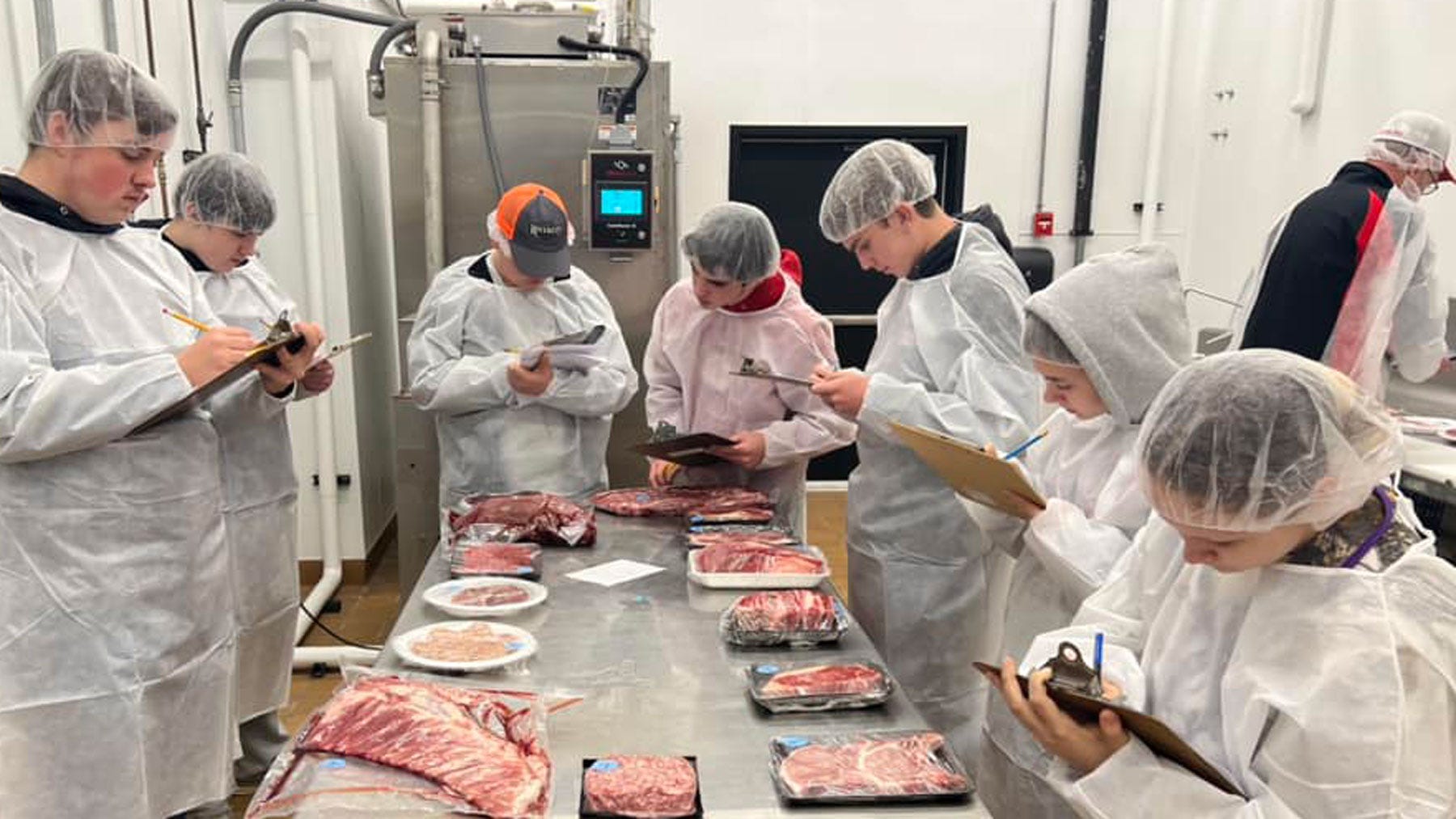 College students dressed in butcher coats and hair nets standing around a table, holding clipboards and rating butcher cuts of meat on the table. College students dressed in butcher coats and hair nets standing around a table, holding clipboards and rating butcher cuts of meat on the table.