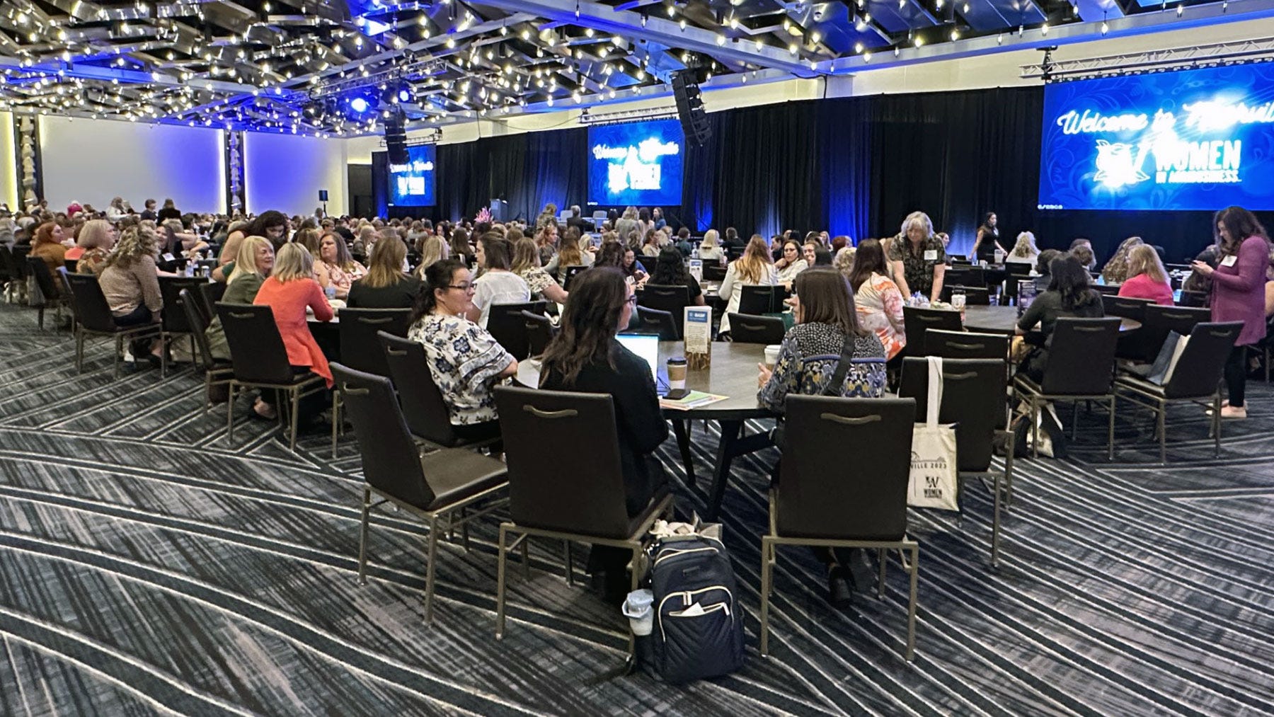 Meeting room full of women attendees at a conference, seated at tables with a stage and three large screens Meeting room full of women attendees at a conference, seated at tables with a stage and three large screens