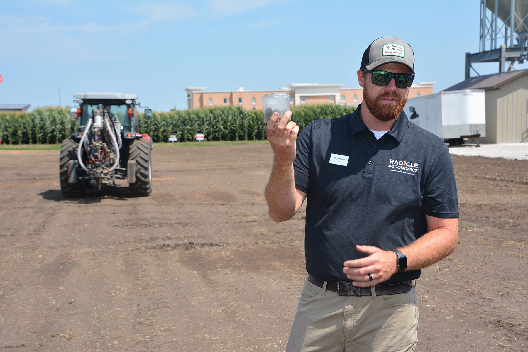 man holds a cup of soil with a tractor and cornfield in the background