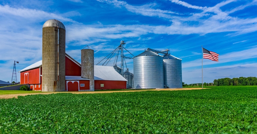 Spring crop fills the foreground leading back to a red barn and rolling hill background with clouds above.