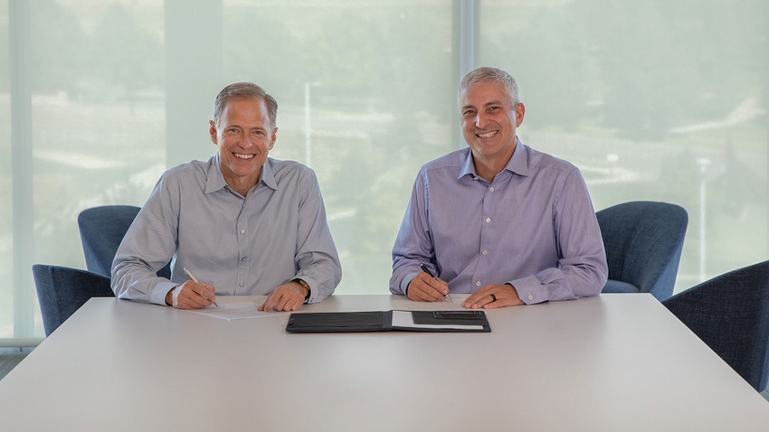 Two men in business casual attire sign papers in an office and smile for a photograph