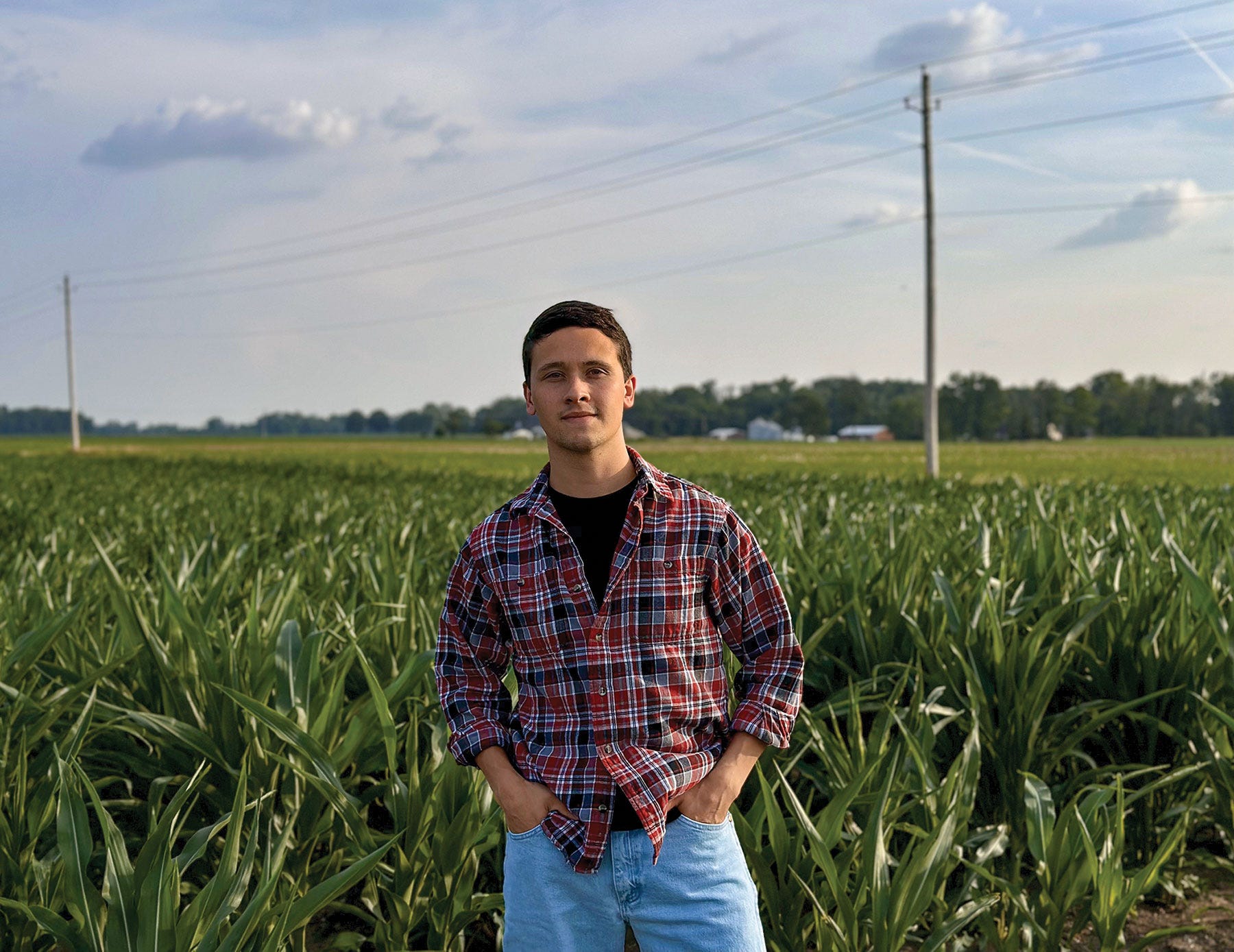 young man in plaid shirt and jeans standing in front of a cornfield