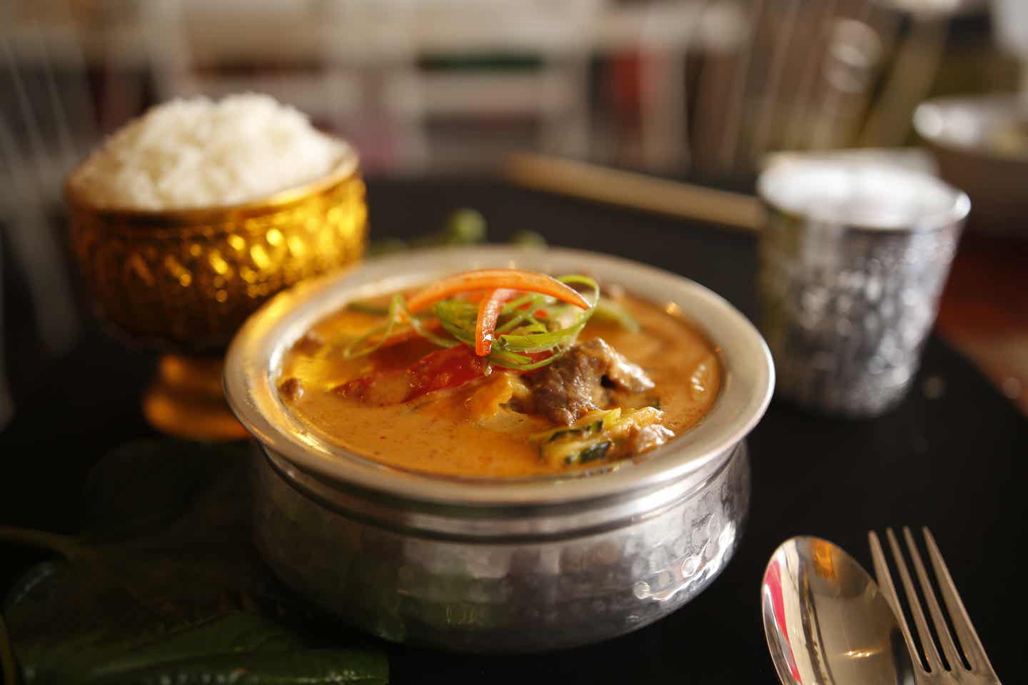 Rotes Thai Curry in einer kunstvollen Metallschüssel. Im Hintergrund: Eine goldfarbene Schale Reis und ein Becher