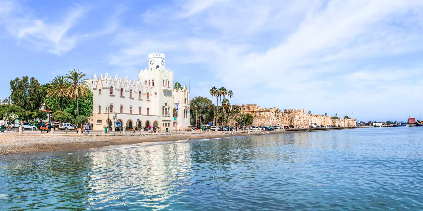 Kos-Tipp: Hafenpromenade der Stadt Kos mit historischem Gebäude, Palmen und ruhigem Meer an der Ägäis