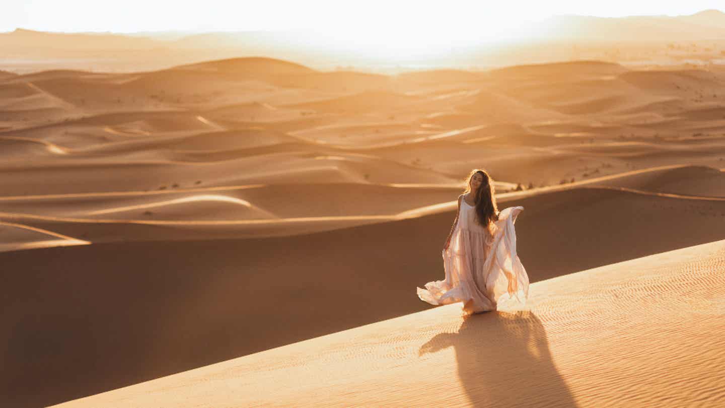 Frau im fließenden Kleid steht in der Sahara bei Sonnenuntergang – Main Character Energy Reise in Marokko