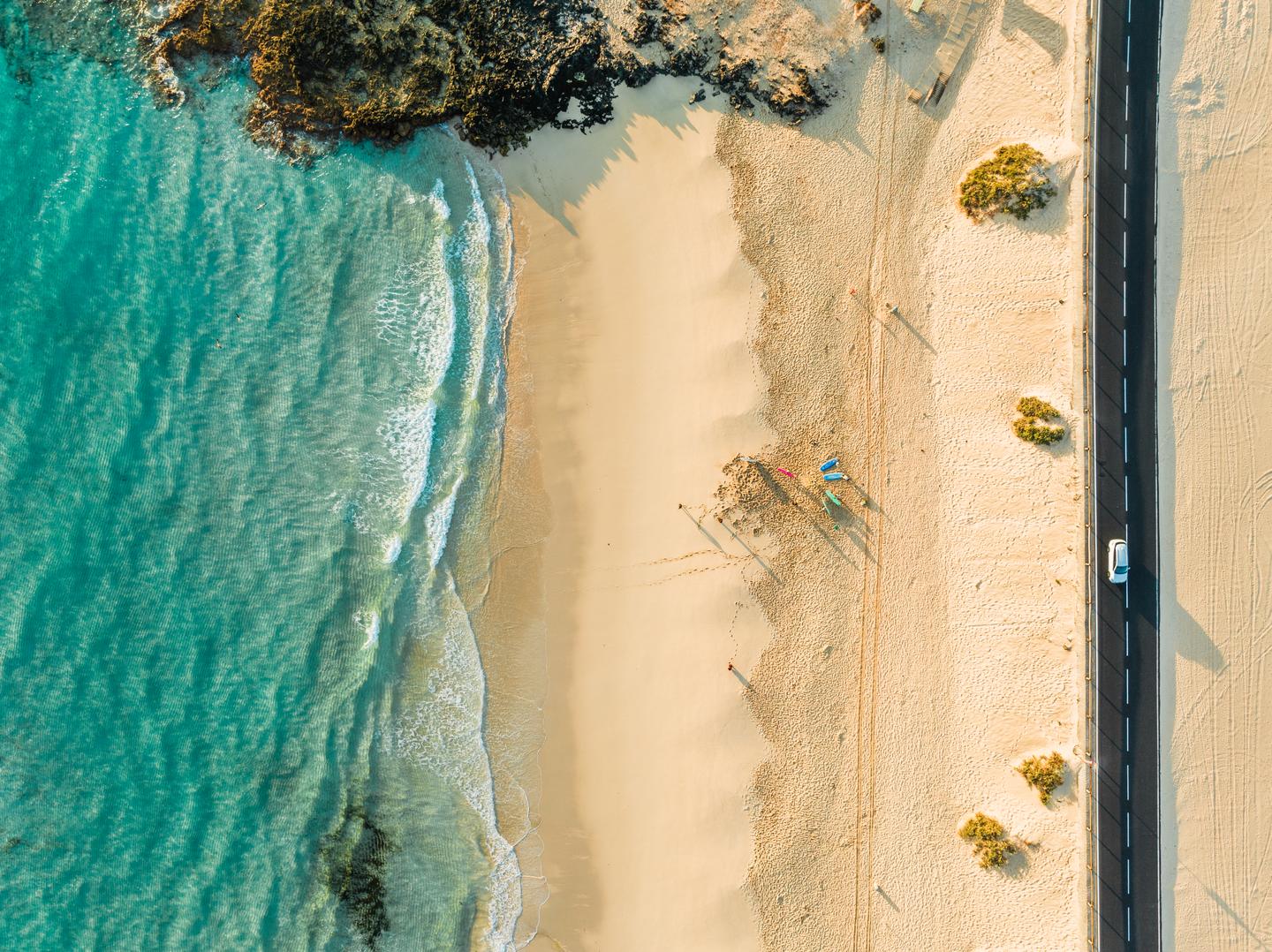 Beste Reisezeit Fuerteventura: Luftaufnahme eines hellen Sandstrands mit türkisblauem Atlantik und Küstenstraße auf Fuerteventura