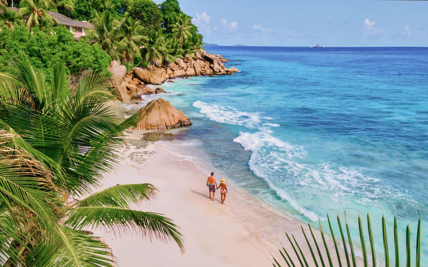 Paar spaziert am weißen Sandstrand von Anse Patates auf La Digue, Seychellen, mit Palmen und türkisblauem Ozean