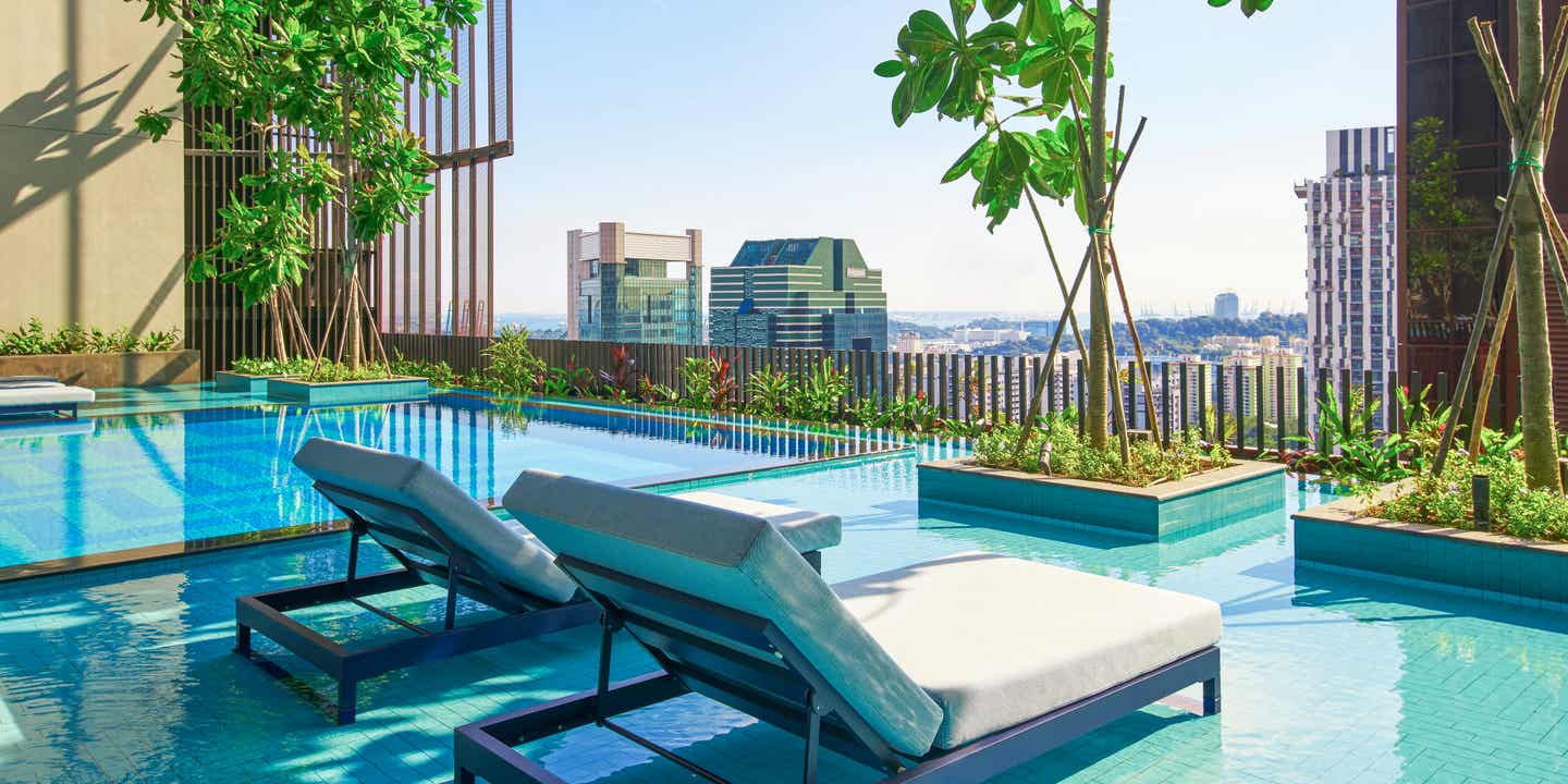 Zwei Liegen am Pool auf einem Hotelbalkon mit Baumbepflanzung und Sicht auf Skyline