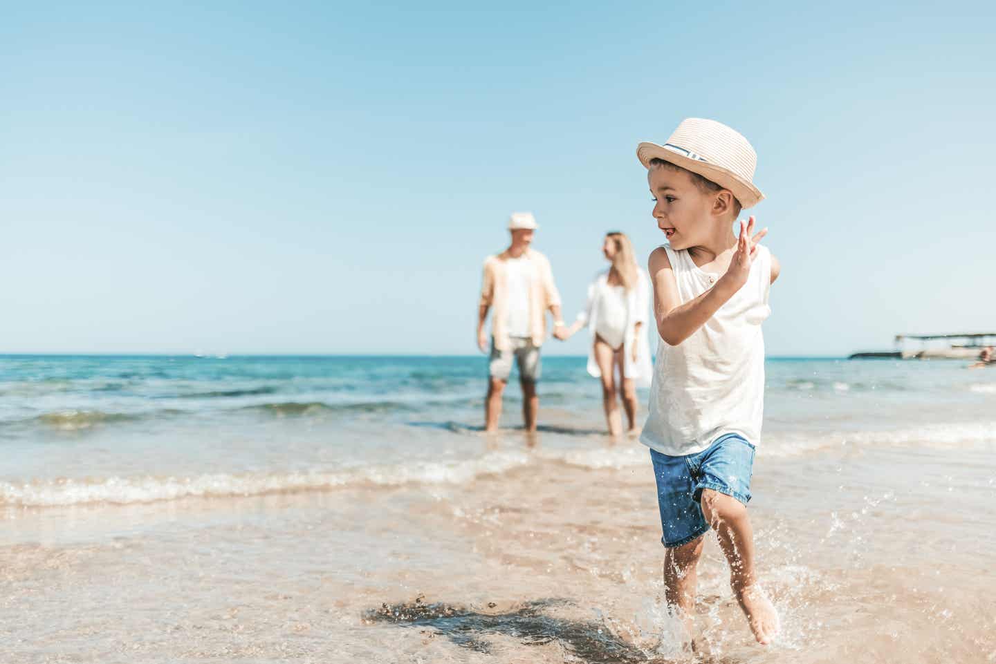 Ein kleiner Junge rennt im Familienurlaub durch das Wasser am Strand, während seine Eltern im Hintergrund Händchen halten