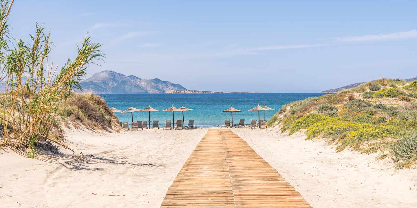 Sandweg durch Dünen zu einem Strand auf Kos mit türkisblauem Meer und Liegen unter Sonnenschirmen