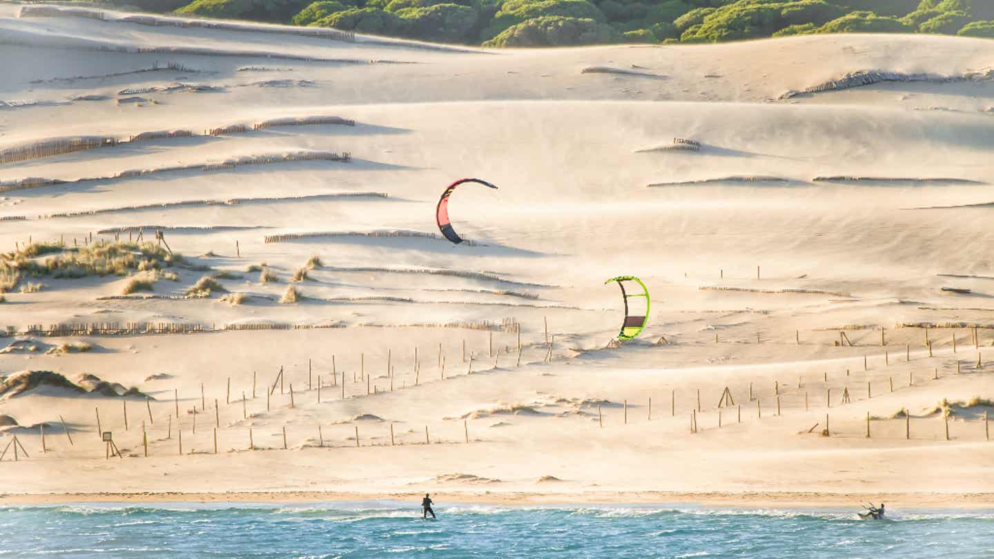 Kitesurfer gleiten vor den Dünen von Tarifa übers Wasser – markante Landschaft zwischen Meer und Windschutz 