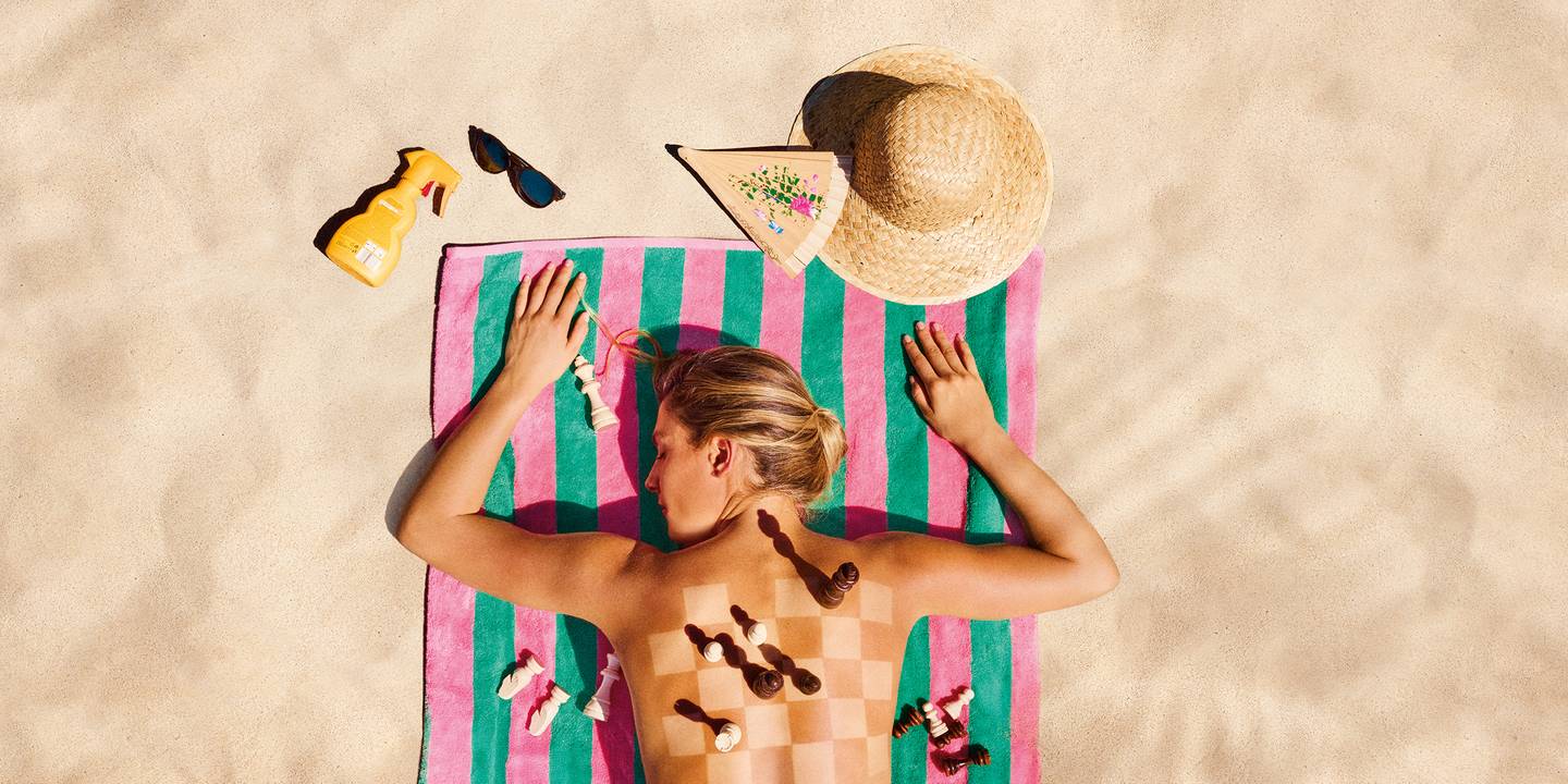 Last Minute 2026: Frau liegt auf einem Strandtuch im Sand, auf ihrem Rücken ist ein Schachbrett mit Figuren. Neben ihr liegen Sonnencreme, Hut, Brille und Fächer