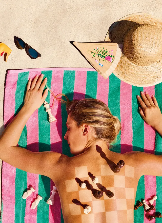 Last Minute in den Urlaub Last Minute 2026: Frau liegt auf einem Strandtuch im Sand, auf ihrem Rücken ist ein Schachbrett mit Figuren. Neben ihr liegen Sonnencreme, Hut, Brille und Fächer