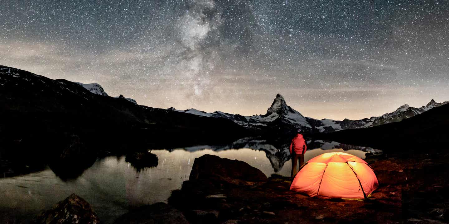 Ein Camper steht vor seinem beleuchteten Zelt und blickt auf den Sternenhimmel; im Hintergrund das Matterhorn