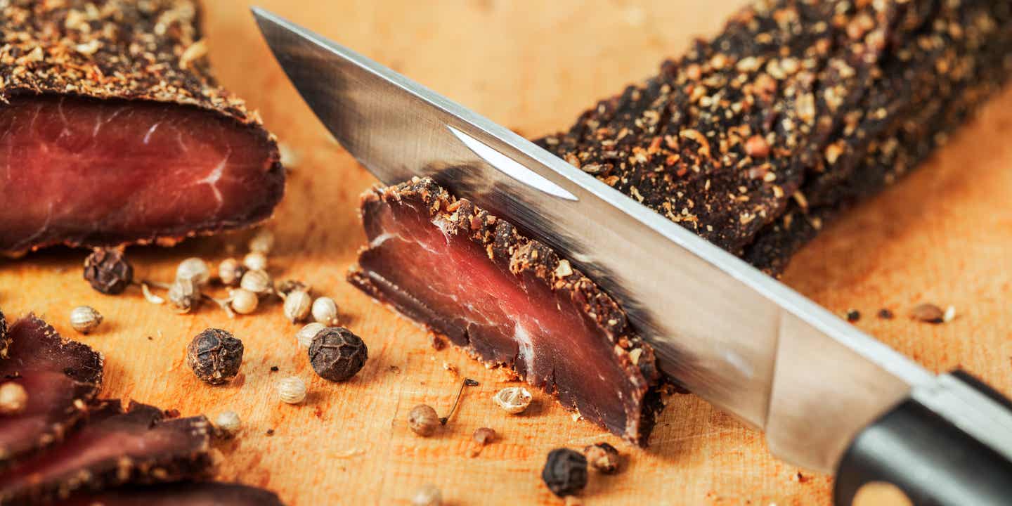 Eine Messerklinge schneidet eine Scheibe von einem schmalen, langen Stück gedörrten Fleisches mit Pfeffer-Kruste, das auf einem Holzbrettchen liegt.