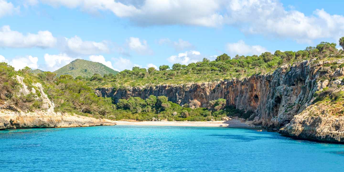 Cala Magraner auf Mallorca mit türkisblauem Wasser, hellem Sandstrand und steilen Felswänden unter blauem Himmel