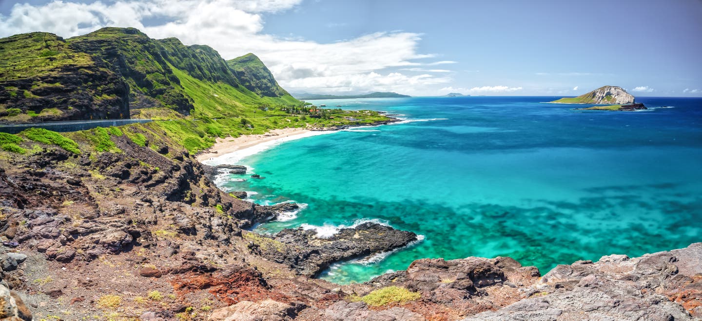 Blick auf den Makapuu Beach, einer der schönsten Hawaii-Strände, mit türkisblauem Wasser vor grün bewachsenen Bergen im Gegenlicht.