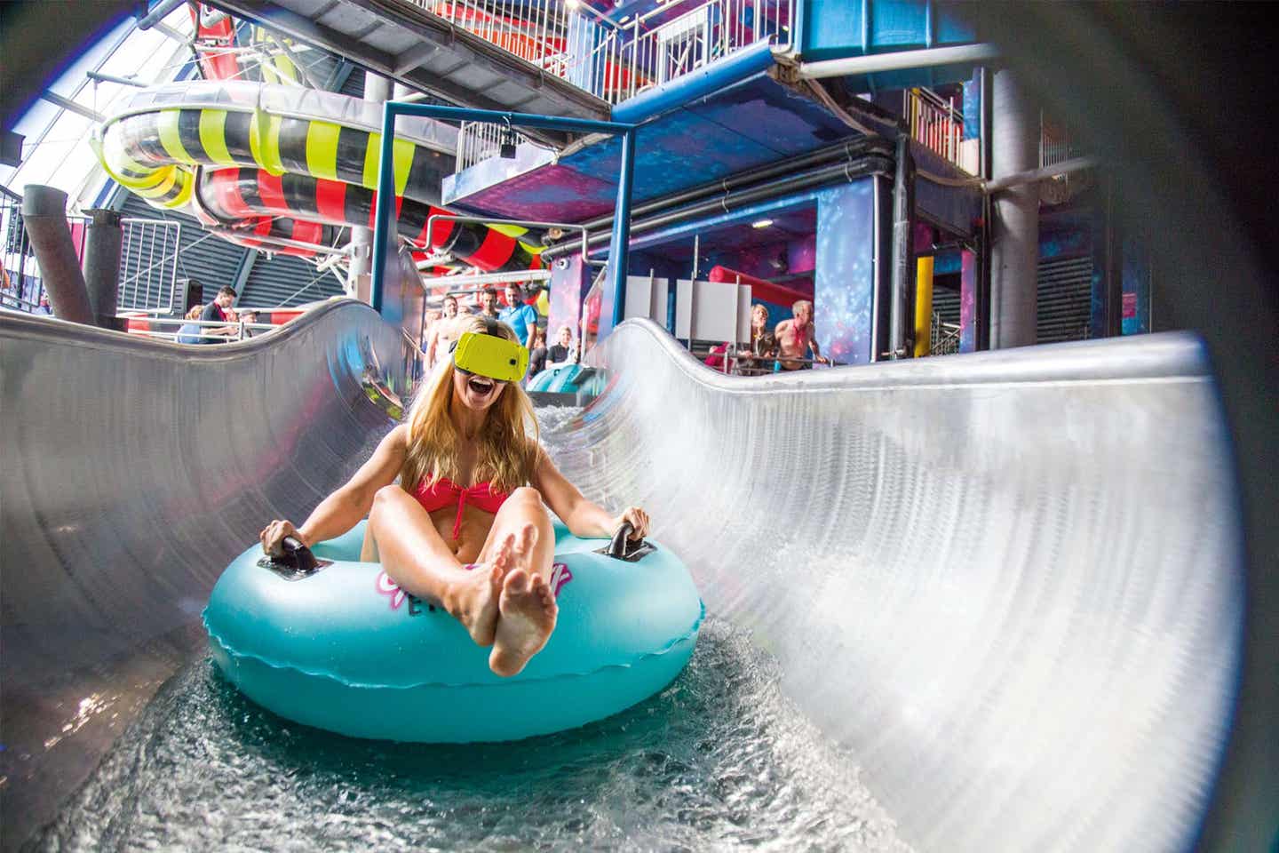 Eine Frau sitzt auf einem Schwimmdonut und fährt mit einer Virtual-Reality-Brille eine Rutsche in der Therme Erding herunter