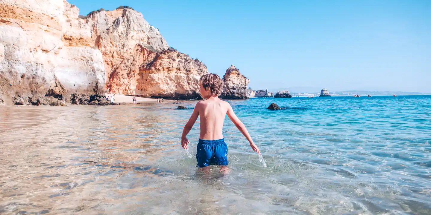 Kinder spielen im flachen Wasser an der Küste von Lagos, Portugal, mit Felsen der Algarve im Hintergrund
