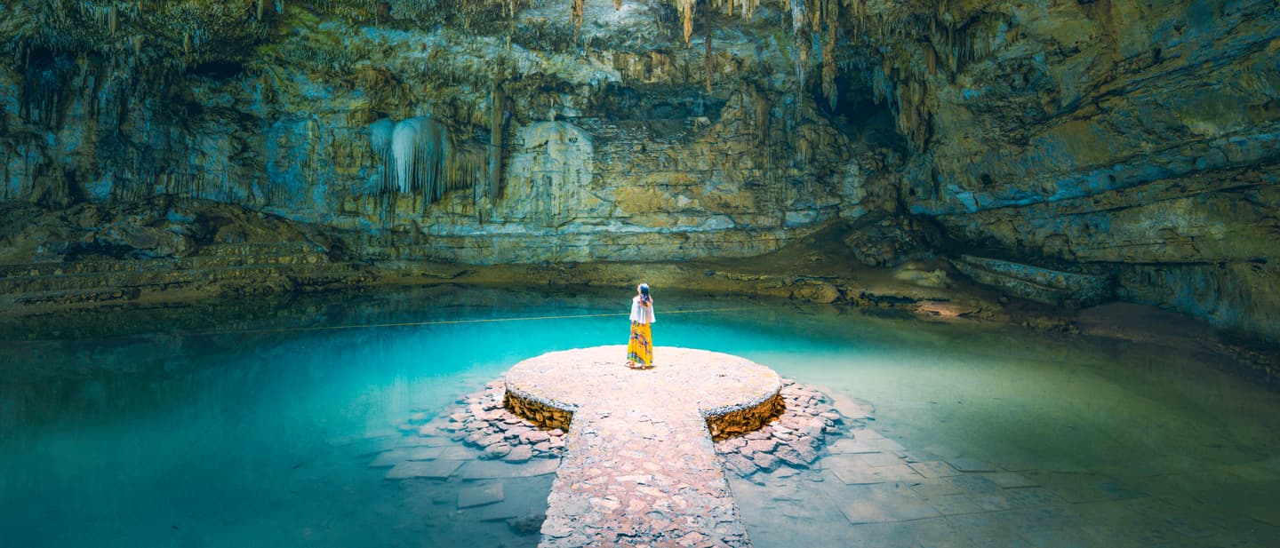 Maßgeschneiderte Reisen: Frau auf einem steinernen Steg in einem von Felsen umgebenen Cenote mit türkisfarbenem Wasser