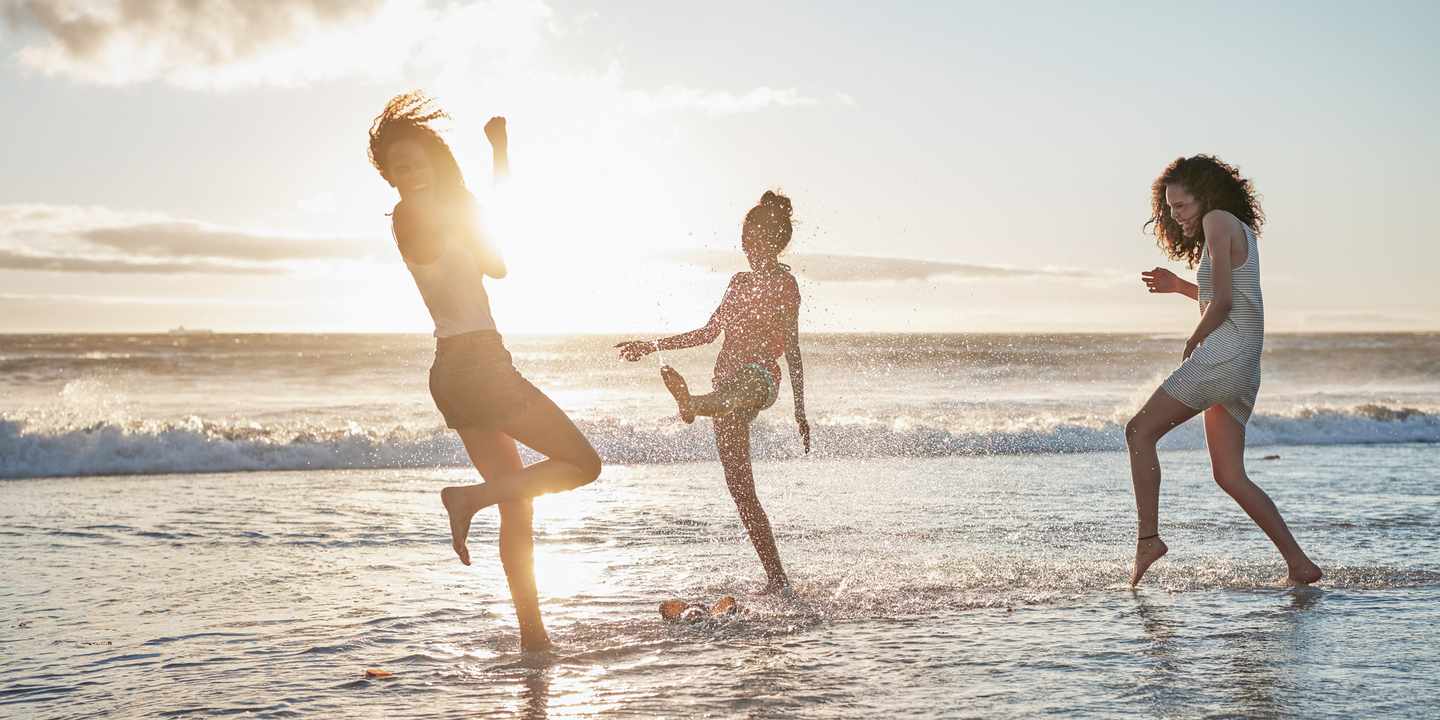 Drei Freundinnen lachen und laufen barfuß durch die Brandung bei Sonnenuntergang während eines Wellnesswochenendes