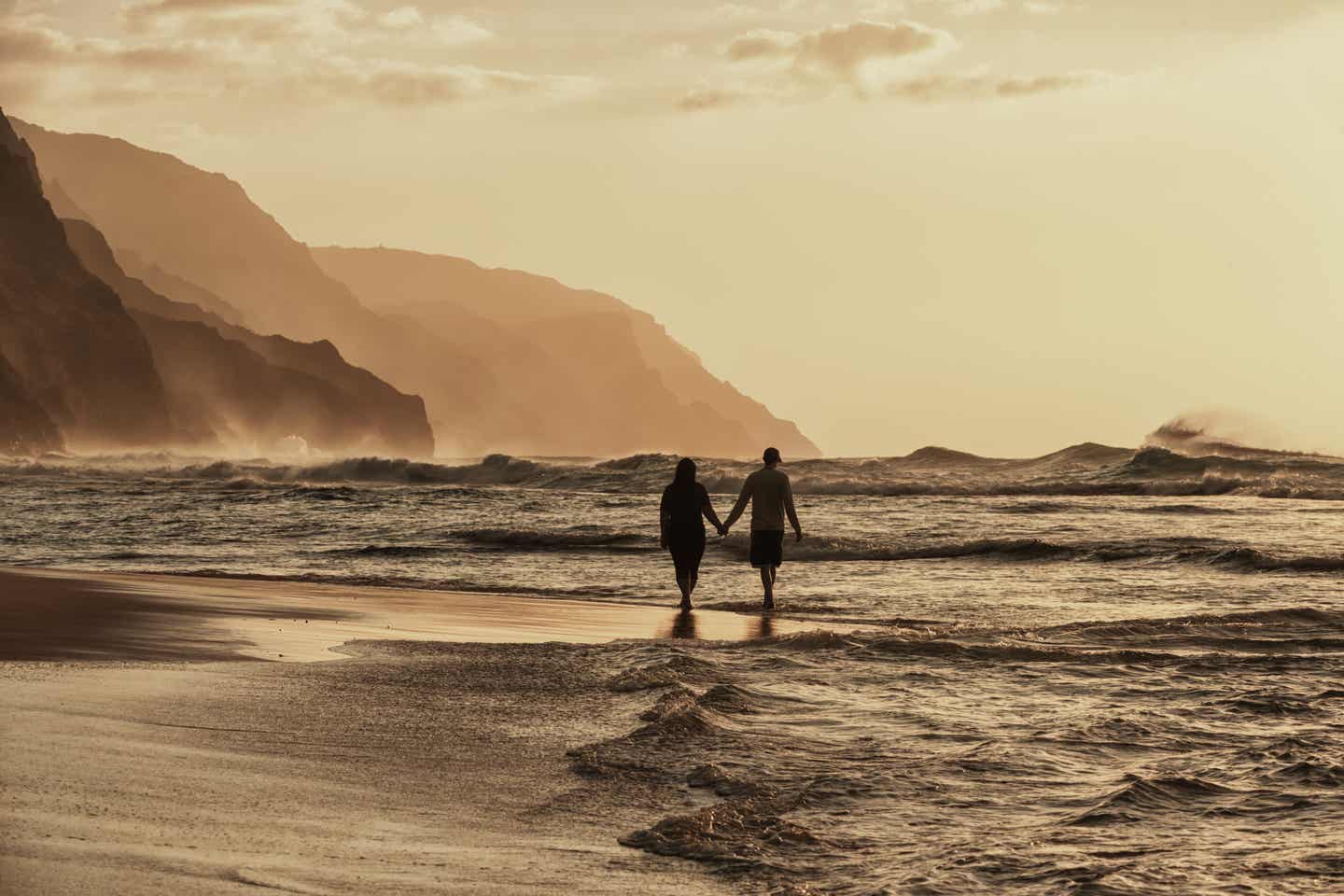 Kauai Urlaub mit DERTOUR. Paar spaziert Hand in Hand bei Sonnenuntergang an einem Strand Hawaiis