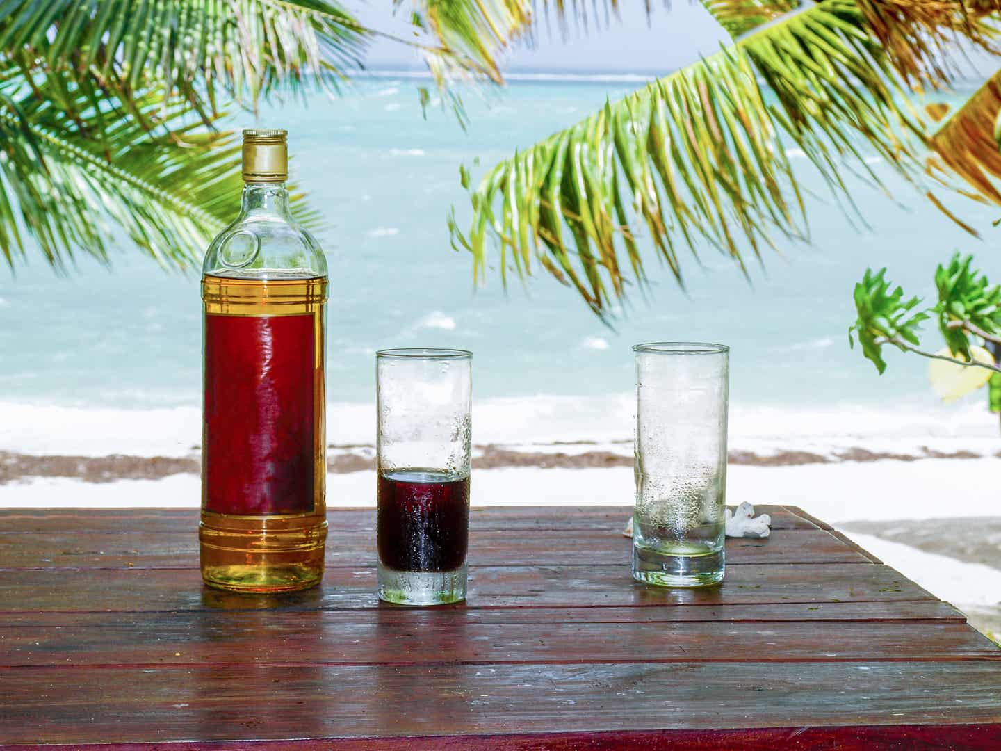 Eine Flasche Rum und ein Glas Cola stehen auf einem Tisch, im Hintergrund befindet sich der Strand