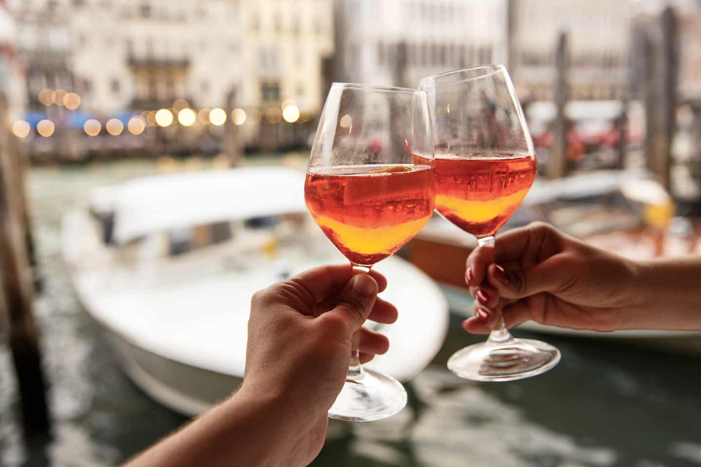 Bibione Urlaub mit DERTOUR. Paar stößt mit Aperol Spritz an. Im Hintergrund unscharf ein Kanal in Venedig