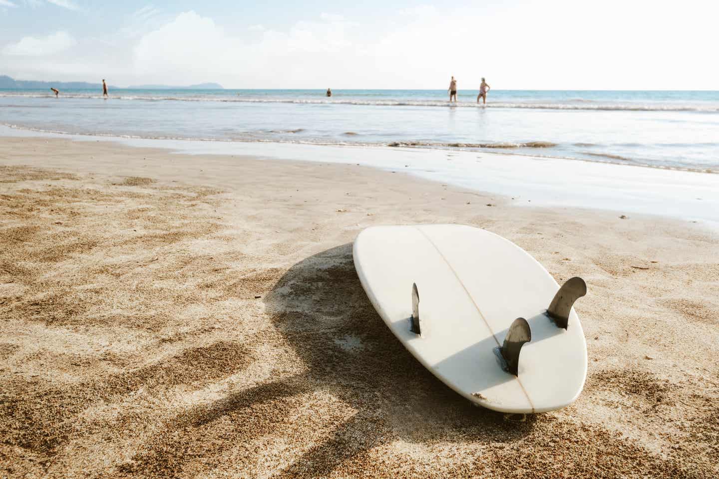 Ein umgedrehtes weißes Surfbrett liegt auf einem Sandstrand, Wellen im Hintergrund