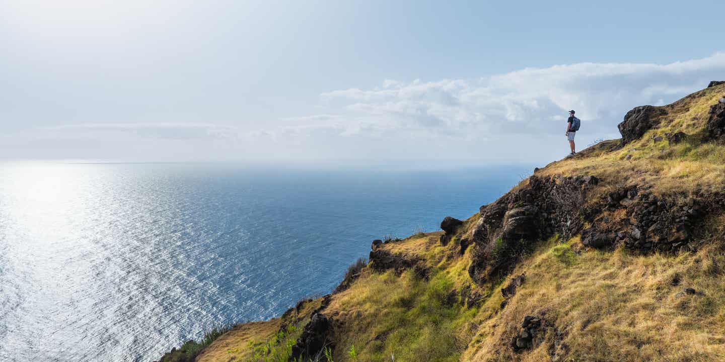 Wanderer steht an einer grün bewachsenen Klippe und schaut auf den blauen Ozean