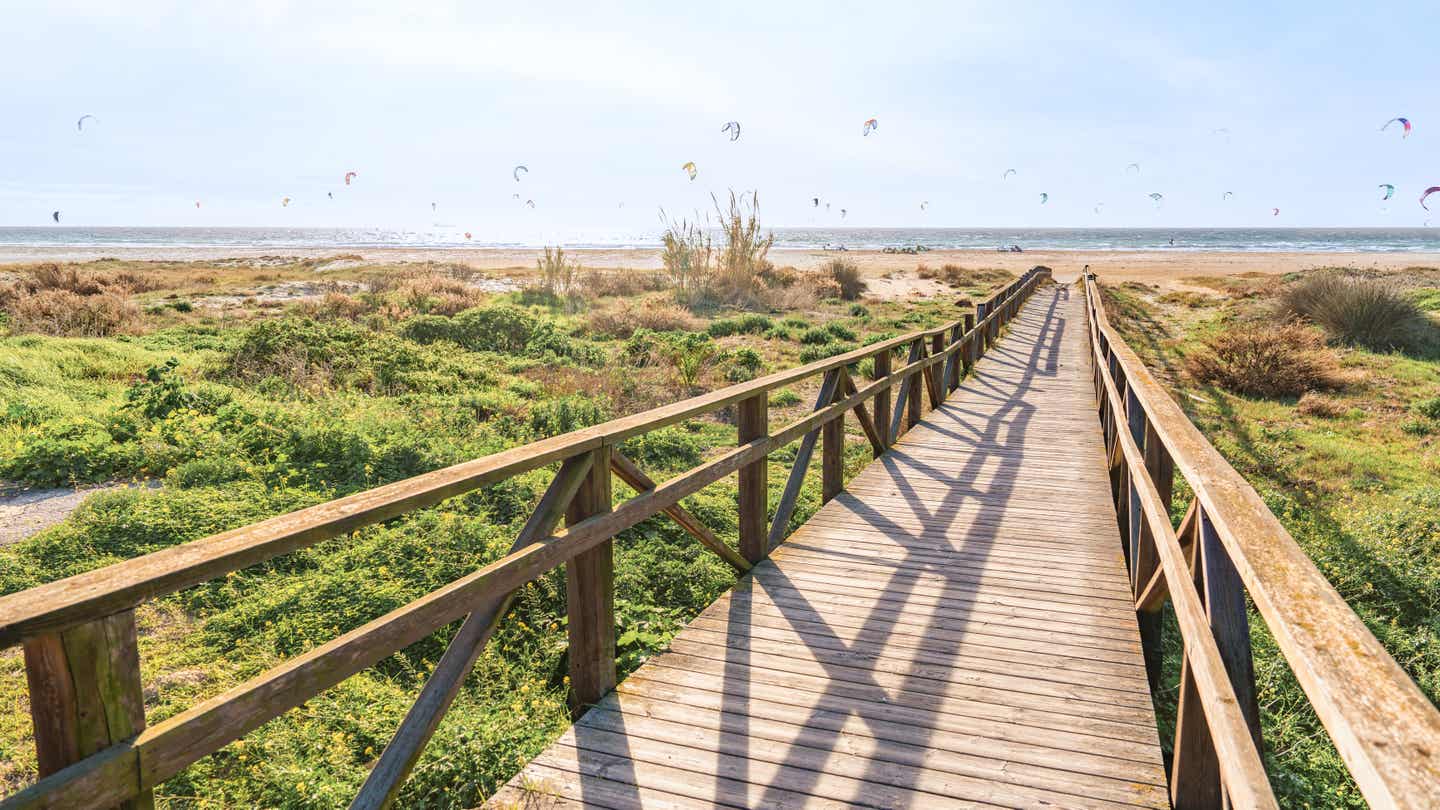 Holzweg führt durch die Dünenlandschaft zum Strand von Tarifa mit bunten Kites am Himmel