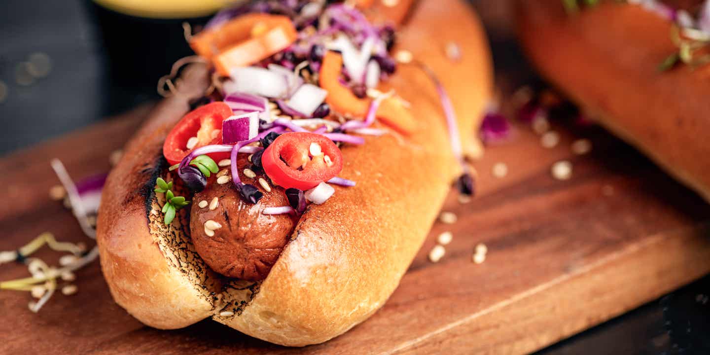 Ein Hotdog mit roten Zwiebeln, Chilis und Sprossen drauf auf einem Holzbrettchen.
