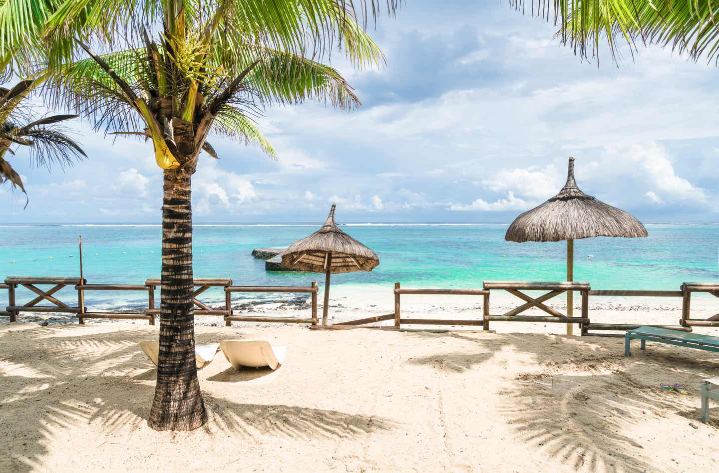 Der mit schattenwerfenden Palmen umzäunte Blue Bay Beach auf Mauritius, mit Sonnenschirmen und türkisfarbenem Wasser im Hintergrund