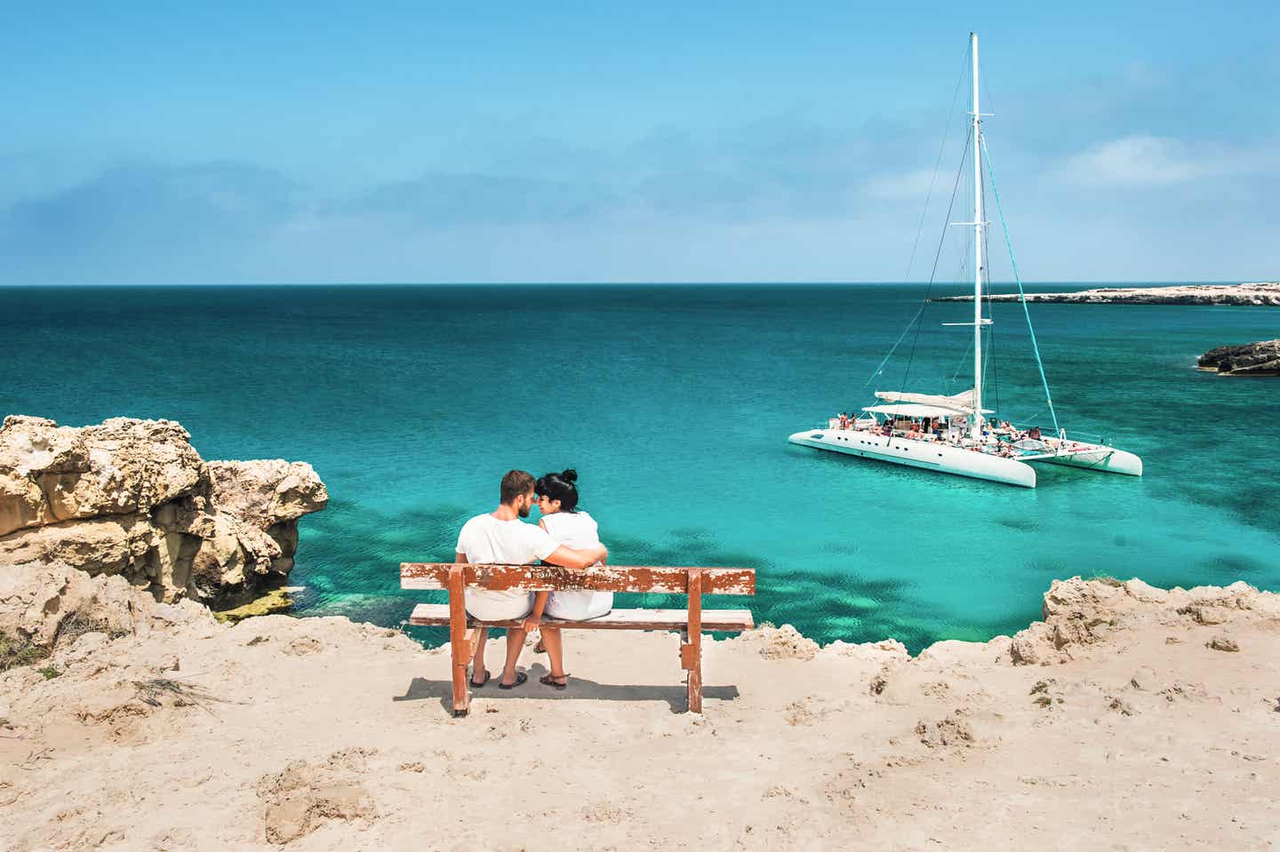 Ein Pärchen sitzt auf einer Bank am türkisblauen Meer mit einem kleinen Boot im Hintergrund