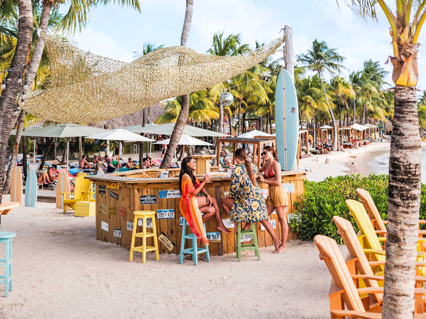 Frauen an Strandbar auf Curacao, sowie bunte Stühle, Palmen und Surfbretter.
