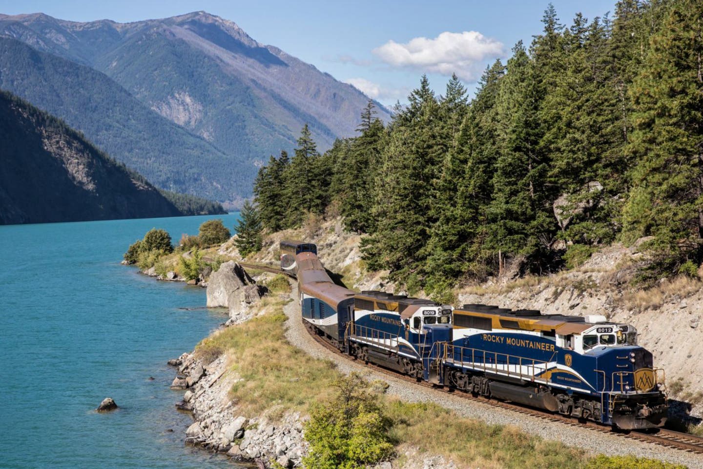 Rocky Mountaineer fährt durch die kanadischen Rocky Mountains mit dicht bewachsenen Wändern auf der linken und einem türkisblauen See auf der rechten Seite