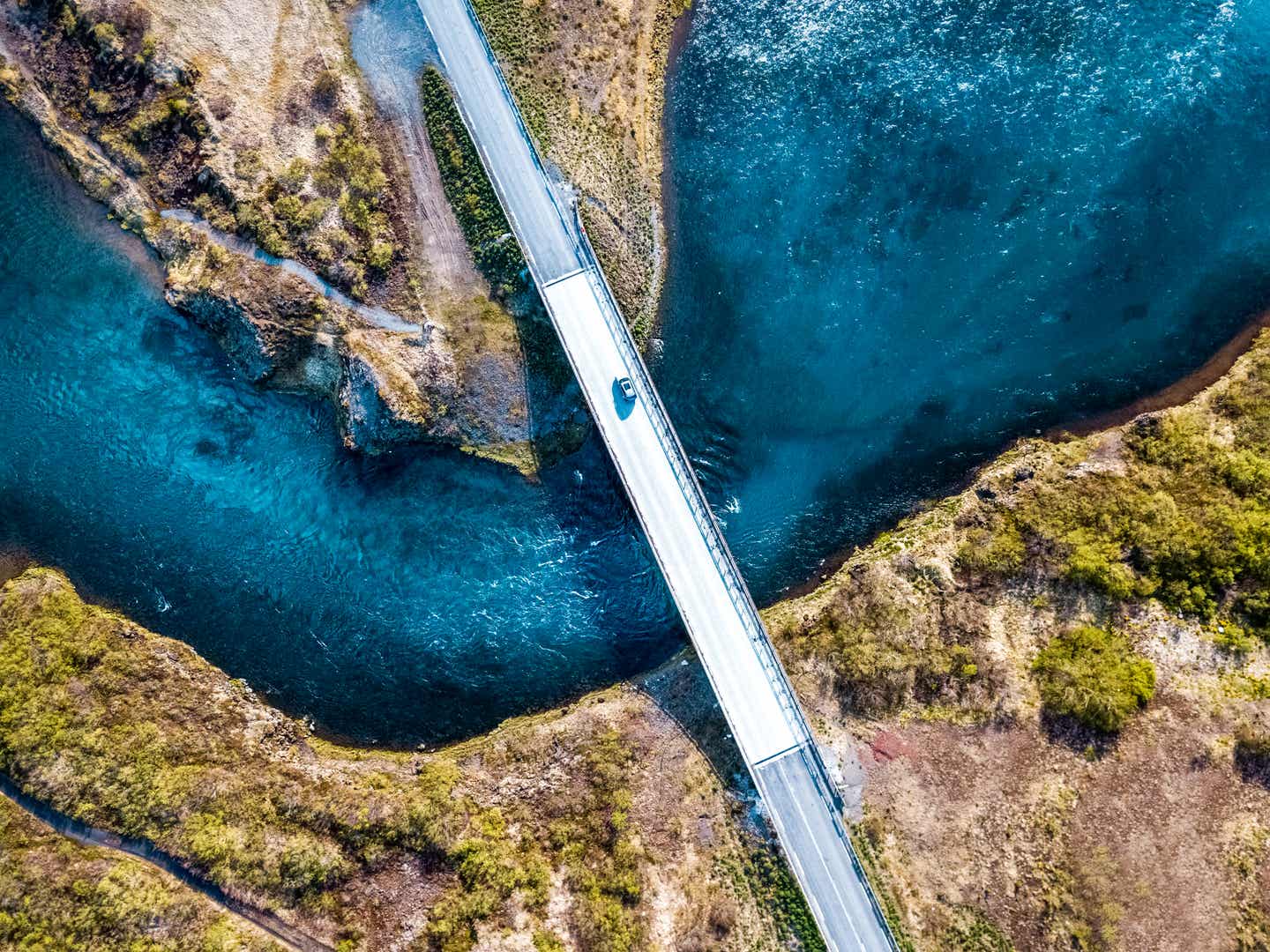 Ein Auto fährt über eine schmale Brücke in Island, darunter ein verzweigter Fluss mit tiefblauem Wasser