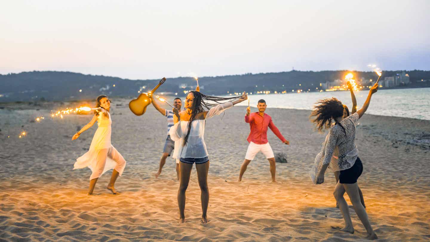 Fünf junge Menschen feiern am Strand mit brennenden Leuchtraketen