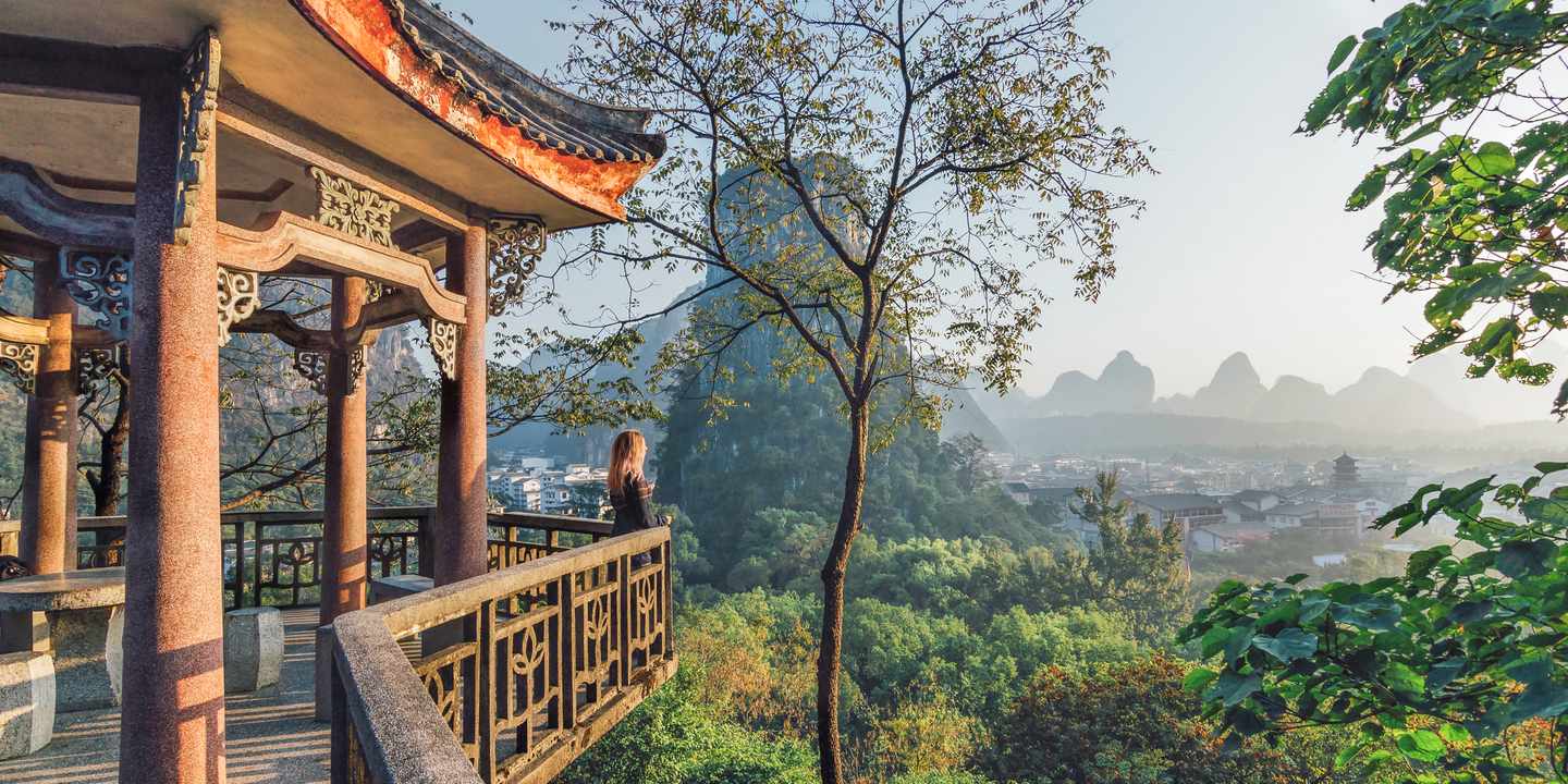 Holzterrasse mit traditionellem chinesischen Dach vor Landschaft mit Karstbergen und einem Dorf im Tal