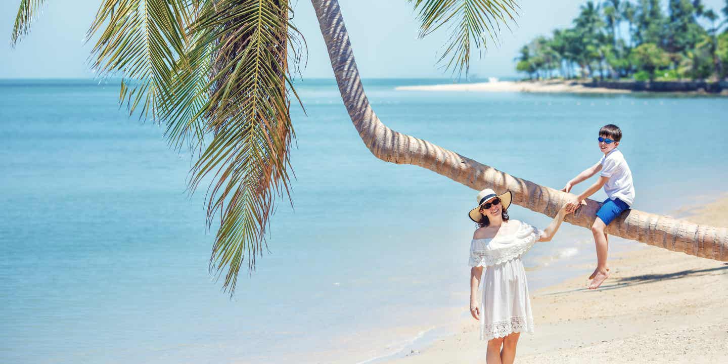 Ein Junge sitzt auf einer schräg wachsenden Palme am Strand, eine Frau in weißem Kleid steht daneben und hält die Palme fest.