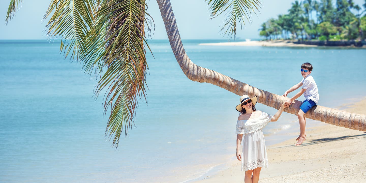 Ein Junge sitzt auf einer schräg wachsenden Palme am Strand, eine Frau in weißem Kleid steht daneben und hält die Palme fest.
